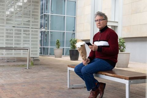Senior man sitting on bench holding bouquet and checking smartphone in modern urban plaza