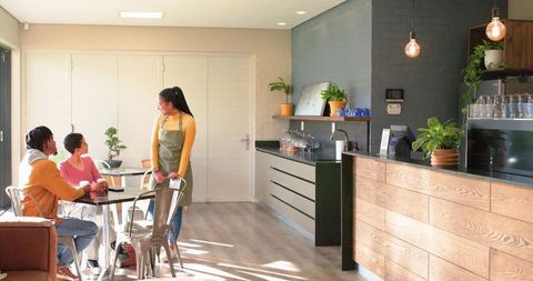 Barista serving two customers at sunlit modern cafe with plants and wooden counter