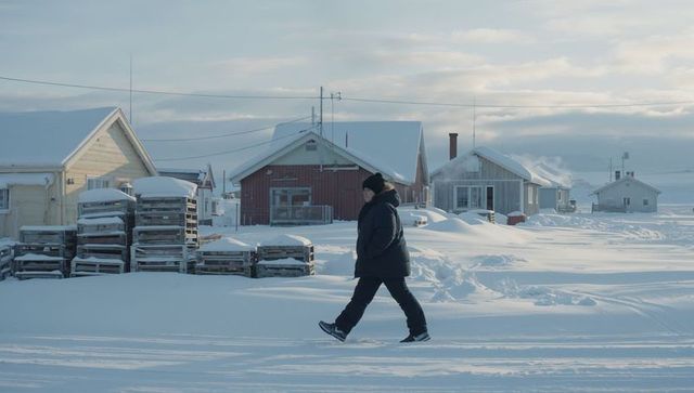 Person walking through snow-covered coastal village with stacked crates, gabled houses
