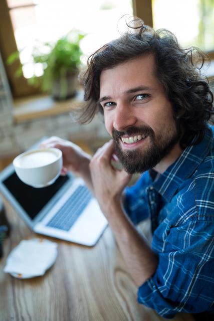 Man sitting in coffee shop holding coffee cup while smiling. The cozy environment and natural light make it perfect for marketing coffee products, promoting coffee shops, or illustrating freelance work in a relaxed setting.