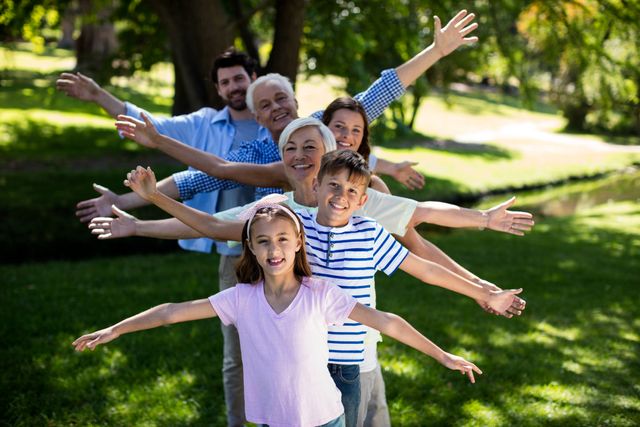 Multi generation family standing in a row with arms outstretched, enjoying a sunny day in the park. Ideal for use in advertisements, family-oriented campaigns, and articles about family bonding, outdoor activities, and healthy lifestyles.