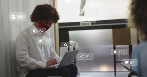 Remote professional typing on laptop in shared lounge wearing red mask with social overlay