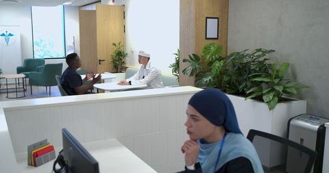 Clinic receptionist wearing navy hijab at reception desk while doctor consulting patient