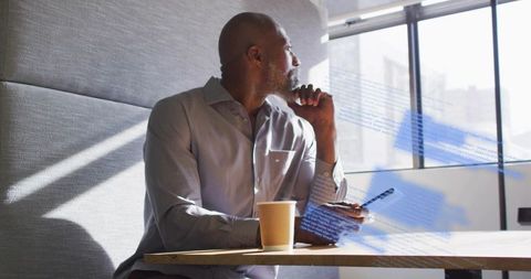 Businessman gazing out window holding smartphone and tablet in modern office booth