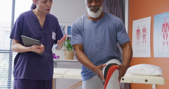 Diverse female physiotherapist and senior male patient holding knee during physical therapy session. Medicine, healthcare, lifestyle and hospital concept.