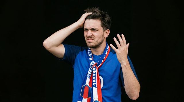 Frustrated soccer fan wearing blue jersey and red white blue scarf expressing confusion