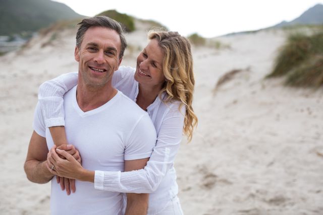 Mature couple standing on a sandy beach, enjoying the moment together. Both individuals are dressed in white, indicating a casual and relaxed atmosphere. Ideal for use in advertisements for beach vacations, lifestyle blogs, articles on relationships and aging, or romantic getaway promotions.