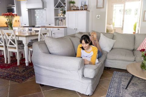Asian woman lounging on gray sectional scrolling smartphone in bright open-plan home