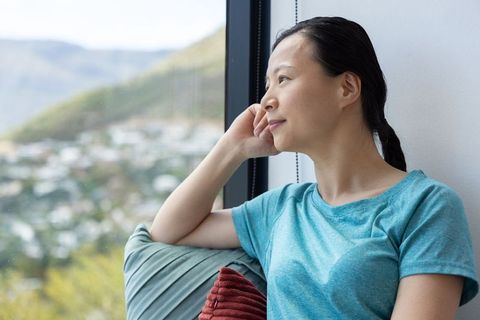 Mature asian woman gazing out window from sofa, contemplative serene wellness mindfulness