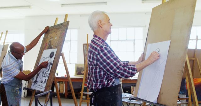 Group of senior artists engaged in sketching at an art studio. They are drawing on large canvases using charcoal and pencils, showcasing their passion for art and creativity. This can be used for articles or advertisements focused on active aging, creative hobbies, senior engagement, and the benefits of art for mental health.