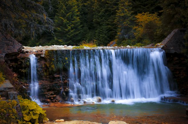 A beautiful waterfall cascades over a ledge into a serene pool of water, surrounded by lush green trees and autumn foliage. This image can be used for nature calendars, travel guides, environmental campaigns, relaxation and meditation content, or as a decorative piece for home and office spaces.