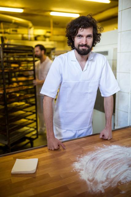 Baker standing at work counter in bakery, smiling and looking at camera. Flour spread on wooden counter, baking racks in background. Ideal for use in articles about baking, small businesses, culinary arts, or professional chefs.