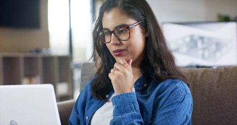 Focused remote professional woman working on laptop from couch with glasses and blue shirt