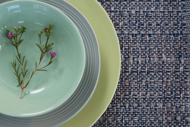 Close-up of an elegant table setting featuring a light green bowl with small pink flowers on a woven placemat. Ideal for use in articles or advertisements about home decor, dining, tableware, or interior design. Perfect for showcasing minimalist and modern dining aesthetics.