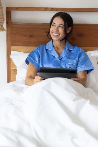 Smiling woman in blue satin pajamas relaxing in bed with tablet for cozy modern morning