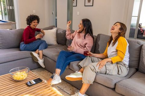 Diverse women laughing on gray sectional sofa during cozy home hangout with drinks