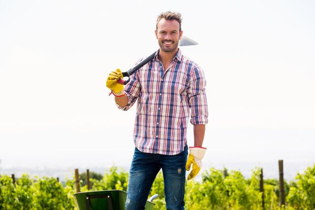 Young man standing in a vineyard, holding a shovel over his shoulder and smiling. He is wearing a plaid shirt, jeans, and gloves, suggesting he is engaged in agricultural or gardening work. The bright, sunny day and the vineyard in the background create a positive and productive atmosphere. This image can be used for topics related to farming, agriculture, rural lifestyle, outdoor work, and gardening.
