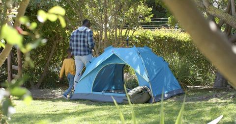 African american father and son walking beside blue camping tent on grassy park lawn