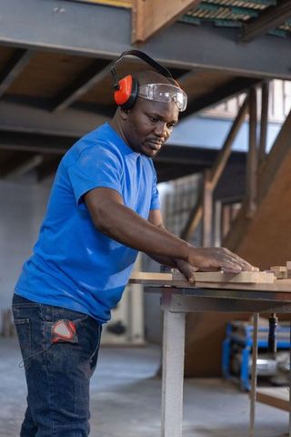 African American carpenter inspecting wooden planks on workbench wearing ear protection