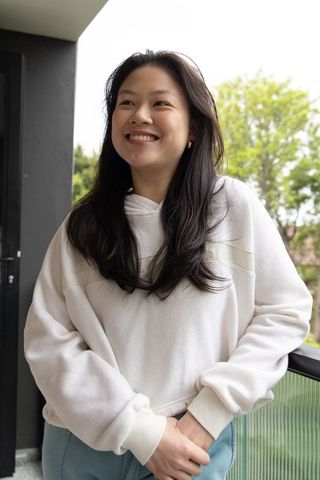 Smiling asian woman leaning on balcony railing wearing cozy hoodie and blue pants