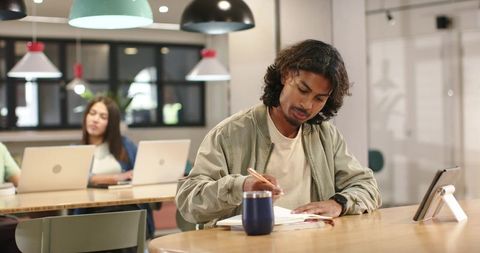 Focused African American Man Writing at Communal Coworking Table with Laptop