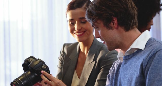 Two colleagues in business attire are using a digital camera, discussing the images they have taken while smiling energetically in a modern office with a relaxed environment. This image can be used to illustrate teamwork, collaboration in a professional setting, creative meetings, or technology in the workplace.
