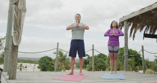 Couple practicing yoga on a wooden deck surrounded by nature. Ideal for promoting holistic wellness, outdoor fitness routines, mindfulness and meditation practices, and healthy lifestyles. Can be used in brochures, online articles, social media campaigns, and wellness programs focused on mature adults or outdoor activities.