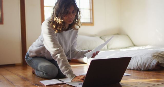 Young woman with curly hair working on laptop seated on wooden floor in cozy bedroom. Closeby notes and papers suggest she's multitasking between her computer and physical notes. Perfect for ads related to freelancing, remote work, studying from home, cozy lifestyle, and work-life balance.