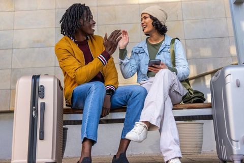 Young travelers high-fiving on station bench with luggage and smartphone, enjoying trip