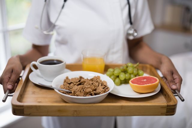 Nurse holding a wooden tray with a healthy breakfast including cereal, coffee, orange juice, grapes, and grapefruit. Ideal for use in healthcare, nutrition, caregiving, and home care contexts. Suitable for illustrating patient care, healthy eating, and medical professional services.