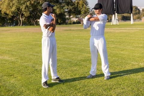 Diverse male cricket teammates stretching arms on sunny field by sight screen warm-up