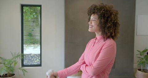 Smiling woman wearing pink zip-up top holding handheld device near window with indoor plants