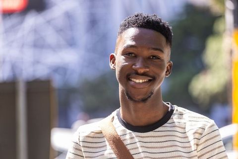 Young black man smiling on sunny city sidewalk wearing striped shirt and crossbody bag