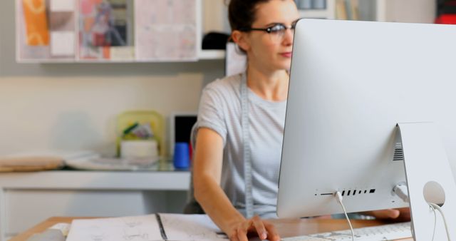 Female architect concentrating on work, using computer in a modern office. She is wearing glasses and navigating through architectural plans on her desk. Ideal for use in contexts related to architecture, design, modern workspaces, female professionals, and creativity in professional environments.