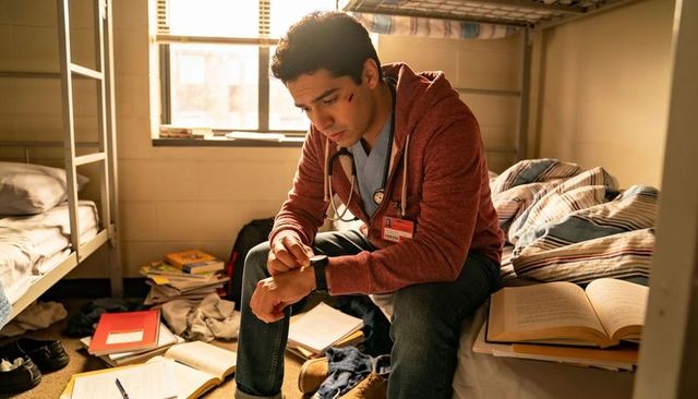 Medical trainee checking watch while sitting on bunk surrounded by textbooks and gear