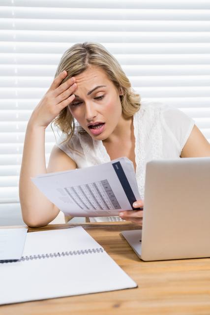 Woman sitting at home office desk, reading a document with a stressed expression. Laptop and paperwork on desk. Useful for illustrating financial stress, work from home challenges, business anxiety, or personal finance management.
