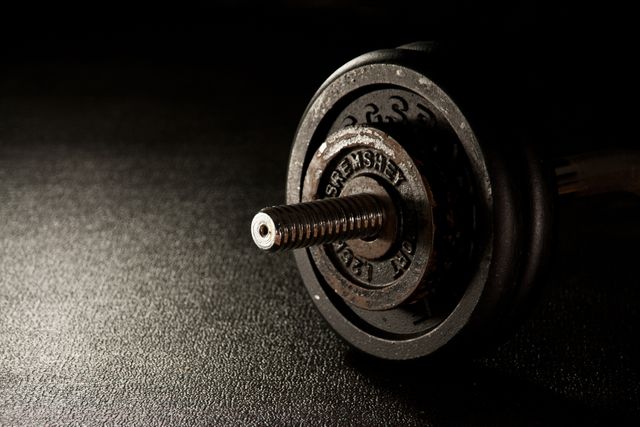 Close-up view of a heavy dumbbell placed on a gym floor with low light highlighting the texture and details of the metal. Ideal for use in fitness-related content, workout guides, gym advertisements, bodybuilding blogs, and strength training promotional material.