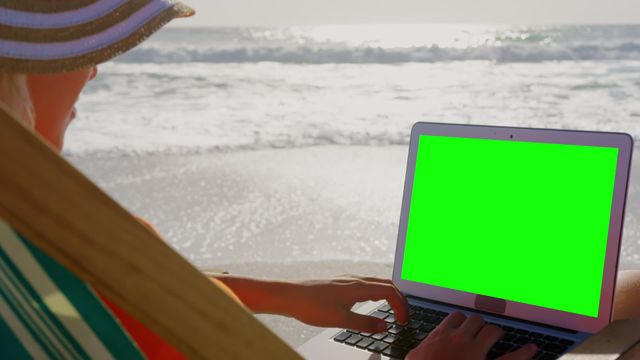 Young woman relaxing on beach, using laptop with green screen. Suitable for themes like remote work, freelance lifestyle, tranquility, and digital nomad. Perfect for promoting tech products or services related to travel and remote working.