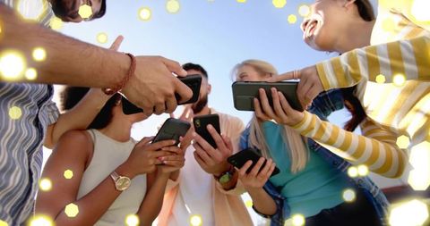 Friends sharing smartphones under blue sky with sunlit hexagonal bokeh and laughter