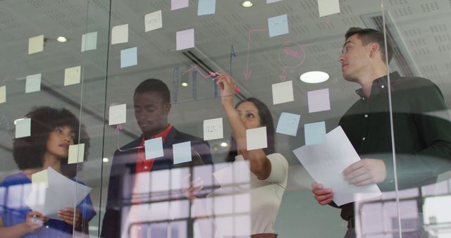 This image shows a diverse group of professionals in an office setting brainstorming together using sticky notes on a glass wall. They appear to be discussing strategies and planning a project. Ideal for use in content related to teamwork, collaboration, corporate culture, business meetings, and project management.