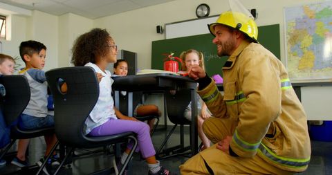 Firefighter kneels beside classroom desk, engaging diverse group of young students in an interactive learning activity about fire safety. Chalkboard and educational map in background create educational atmosphere, perfect for use in promotional materials, educational websites, or community safety awareness campaigns. Emphasizes importance of learning about fire safety from experts at a young age, encouraging future preparedness and awareness.