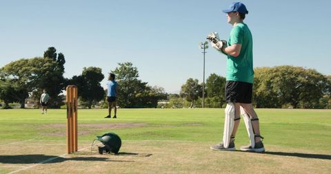 Young batsman standing at wicket on sunny cricket field with helmet, stumps and pads