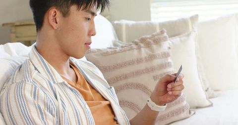 Young asian man checking card on sofa in cozy living room wearing striped overshirt