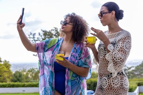 Two women posing for selfie on terrace in swimwear and crochet cover-ups holding yellow drinks