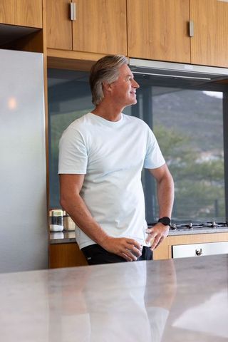 Mature man leaning on marble counter wearing blue tee and smartwatch in modern kitchen