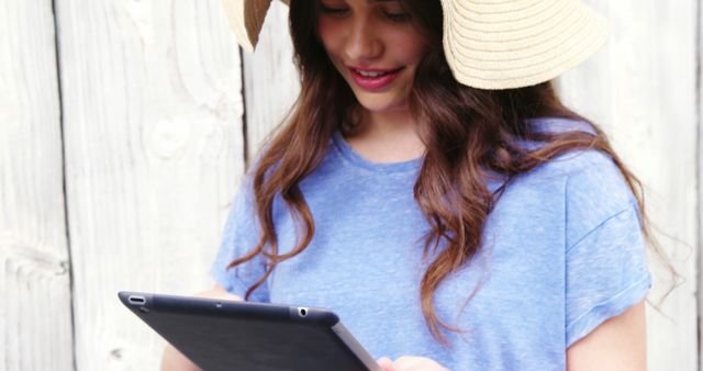 Young woman with long brown hair smiling while using a tablet outside. She is wearing a wide-brimmed sun hat and a casual blue shirt. This image can be used for technology, lifestyle, leisure, and outdoor activities content. It is suitable for articles, blogs, websites, and marketing materials highlighting the use of modern technology in everyday life.