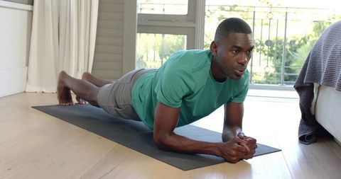 African American man performing forearm plank on yoga mat in bright minimalist bedroom