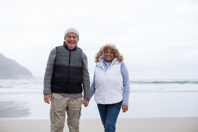 Senior couple enjoying a walk on the beach, holding hands and smiling. They are dressed in winter clothing, indicating a cool day by the ocean. Ideal for use in advertisements, retirement planning brochures, lifestyle blogs, and articles about active aging and relationships.