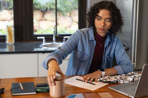 Non-binary creative reaching for coffee while reviewing sketches at sunlit home workstation