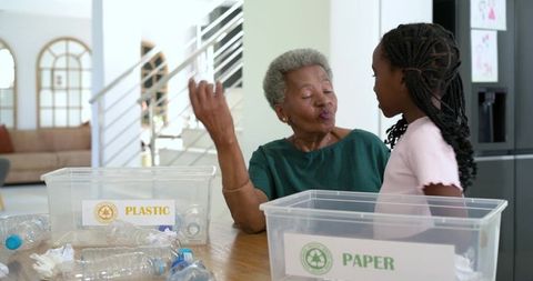 African senior woman teaching granddaughter recycling with labeled plastic and paper bins